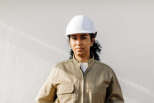 Portrait of a hispanic female electrician standing against white wall. Woman wearing khaki uniform and hardhat looking at camera